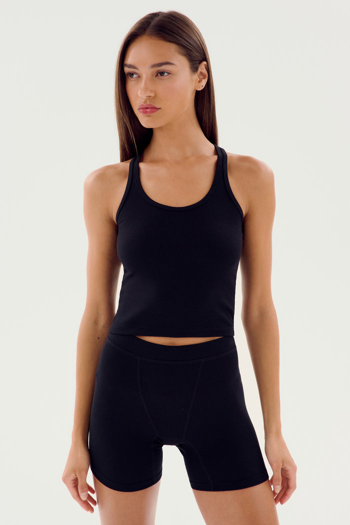 Woman wearing the SPLITS59 Ashby Rib Crop, a fitted black tank top, paired with black high-waisted shorts, standing against a plain light background.
