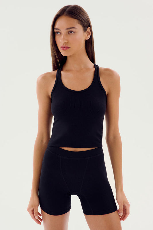 Woman wearing the SPLITS59 Ashby Rib Crop, a fitted black tank top, paired with black high-waisted shorts, standing against a plain light background.