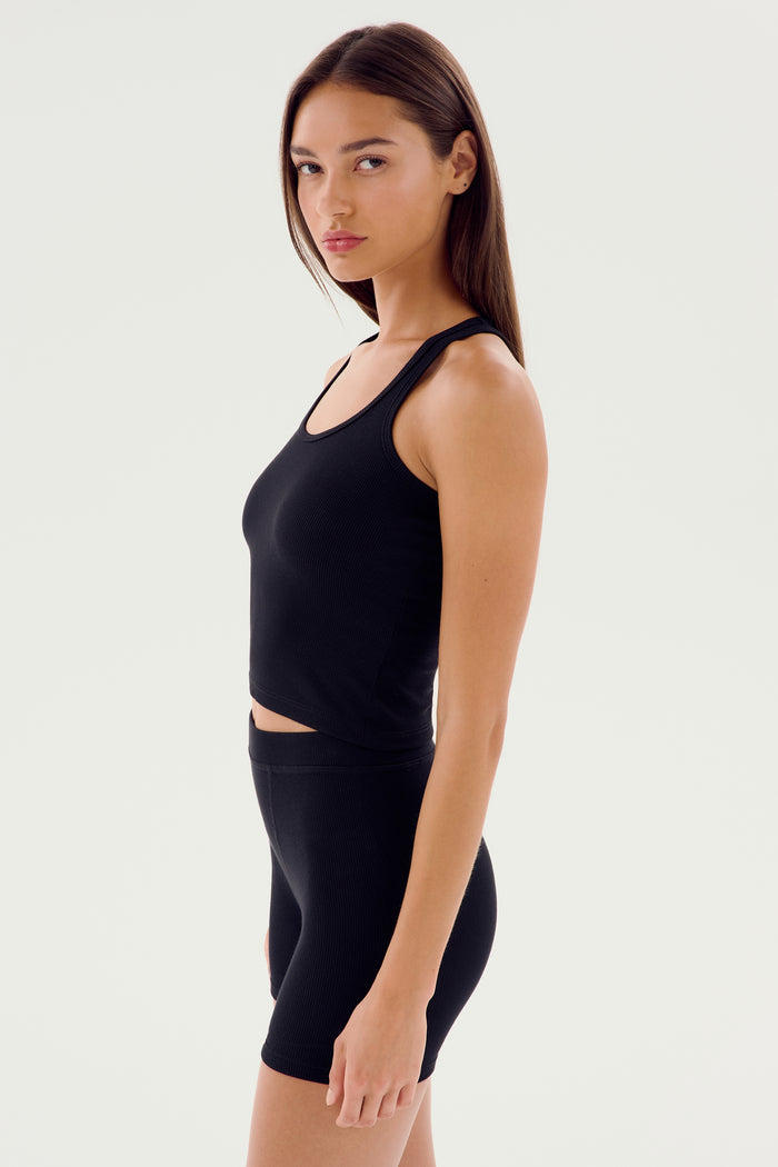 A person with long brown hair wearing the Ashby Rib Crop - Black from SPLITS59 and black shorts, made from baby rib fabric, stands facing slightly to the side against a plain white background. This outfit is perfect for intense gym workouts or casual outings.
