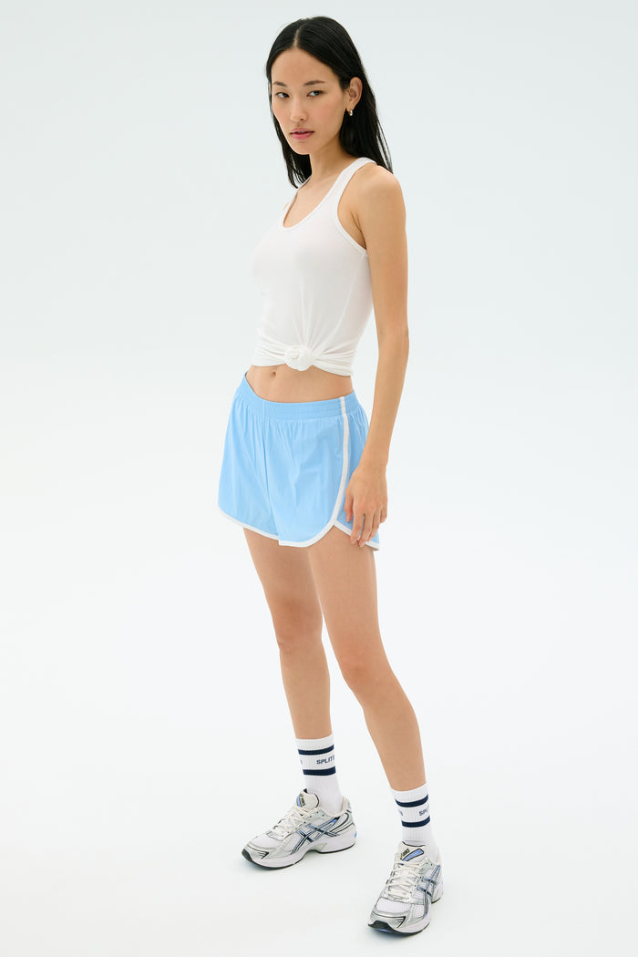 A woman wears the Splits59 Ashby Rib Tank in white, paired with light blue athletic shorts, striped white socks, and running shoes, standing against a plain light background.