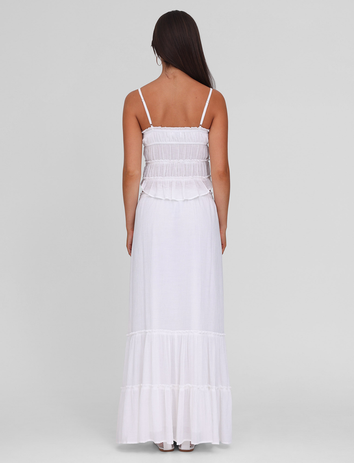 A woman with long dark hair, seen from behind, wears the TIGERMIST ASHLYNN MAXI SKIRT in white. The sleeveless skirt has thin straps, a ruffled tiered design, elasticated waist, and is set against a plain light gray background.