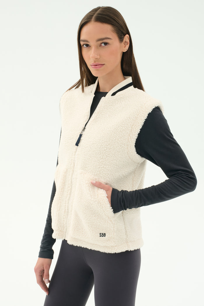 A woman wears the SPLITS59 Aster Sherpa Vest - Creme over a black long-sleeve top and leggings, standing against a plain white background, suited for cool weather workouts.