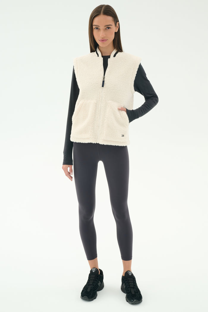 A woman wears the SPLITS59 Aster Sherpa Vest - Creme, a cozy layer for cool weather workouts, over a black long-sleeve top, leggings, and sneakers, standing against a plain white background.