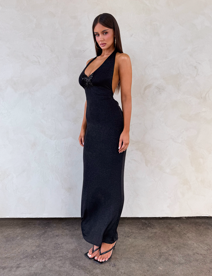 A woman with long dark hair stands against a textured white wall, wearing the TIGERMIST ASTORIA MAXI DRESS in black with a deep neckline and black strappy sandals, facing slightly to the side and looking at the camera.