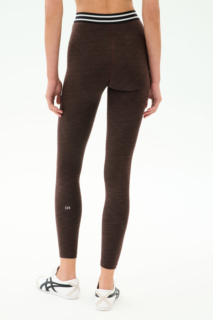A person wears SPLITS59's Bailey High Waist Active Rib 7/8 leggings in Heather Dark Chocolate, featuring a black and white striped waistband and white sneakers, shown from the back—ideal for gym workouts.
