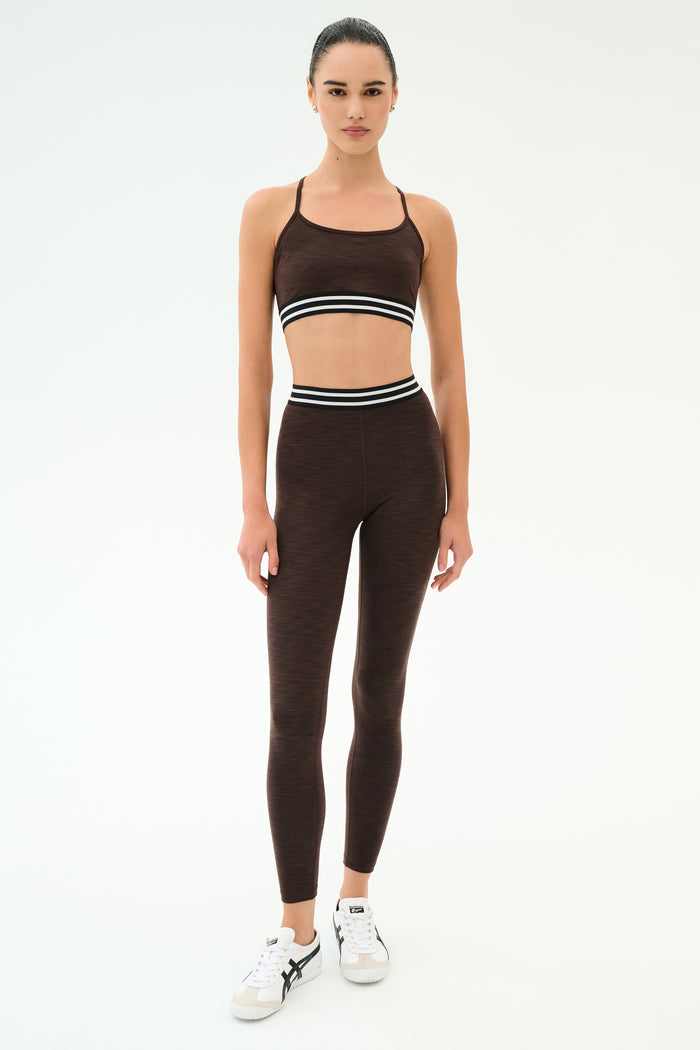 A woman stands against a plain background wearing the SPLITS59 Bailey High Waist Active Rib 7/8 leggings in Heather Dark Chocolate, paired with a matching rib sports bra and white sneakers—ideal for gym workouts.