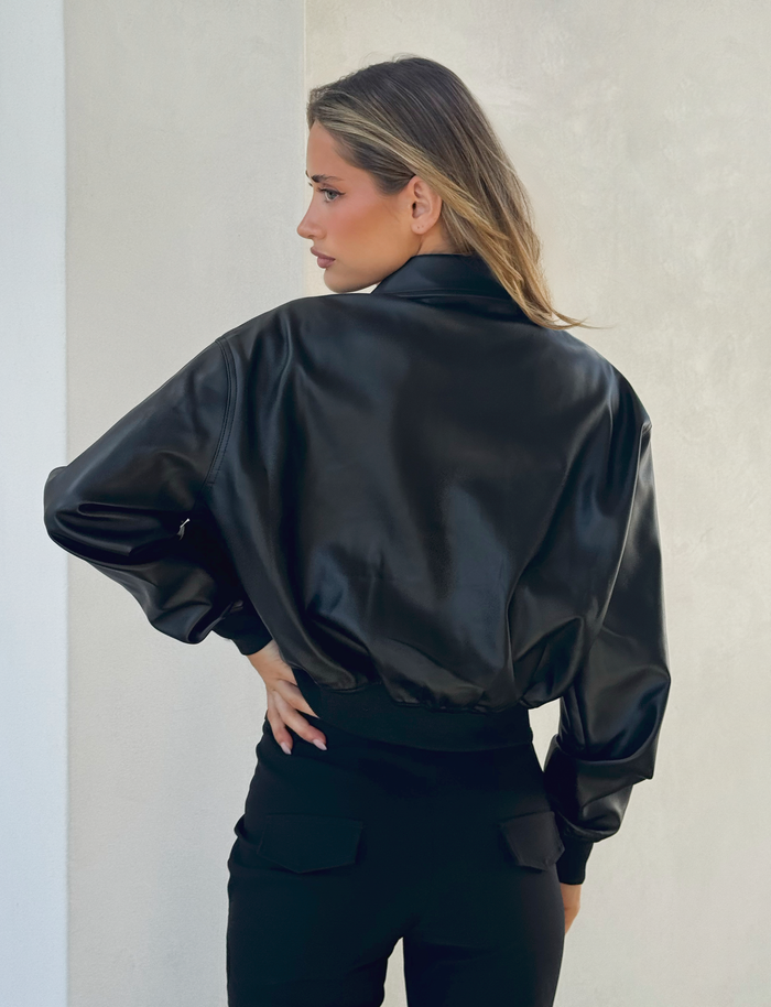 A woman with long, light brown hair stands with her back to the camera, wearing the TIGERMIST BEREN JACKET - BLACK, a faux leather bomber with a collared neck, paired with black pants, against a light-colored wall.
