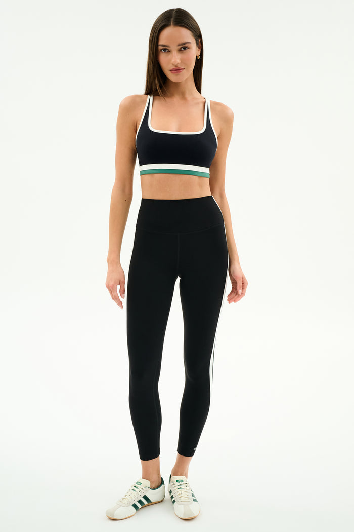 A woman stands facing forward in SPLITS59 athletic wear: a black sports bra with white and green accents, Bianca High Waist Rigor 7/8 leggings in Black/White, and white sneakers with green stripes.