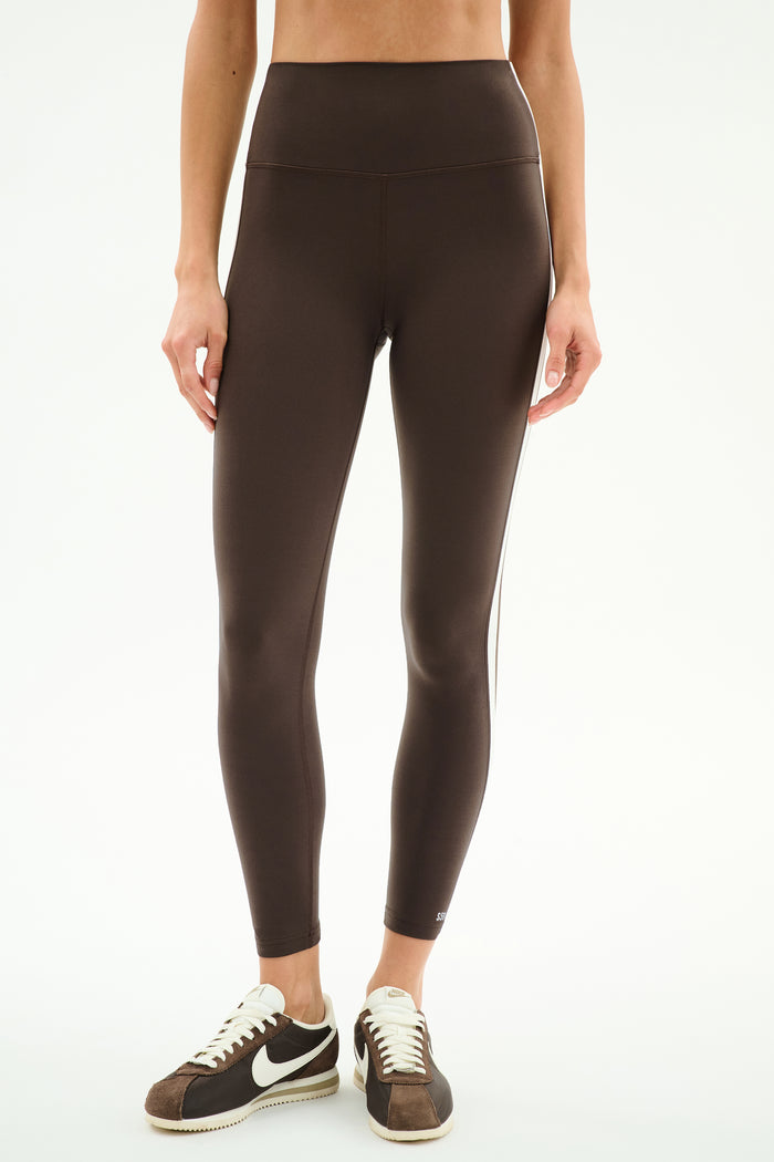 A person wears SPLITS59 Bianca High Waist Rigor 7/8 leggings in Dark Chocolate/White with brown-and-white sneakers, standing against a plain white background.