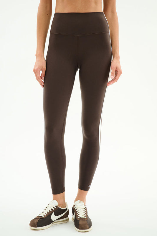 A person wears SPLITS59 Bianca High Waist Rigor 7/8 leggings in Dark Chocolate/White with brown-and-white sneakers, standing against a plain white background.