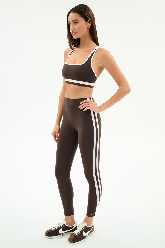 A woman wears the SPLITS59 Bianca High Waist Rigor 7/8 leggings in Dark Chocolate/White, featuring white side stripes and performance fabric, paired with a matching sports bra and black-and-white sneakers.