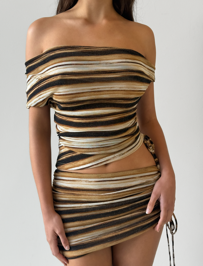 A woman wears the TIGERMIST BRAELYN TOP in brown stripe, featuring form-fitting lurex fabric with horizontal brown, gold, black, and cream stripes. The off-shoulder style highlights her neckline as she stands against a light background, hand on her hip.