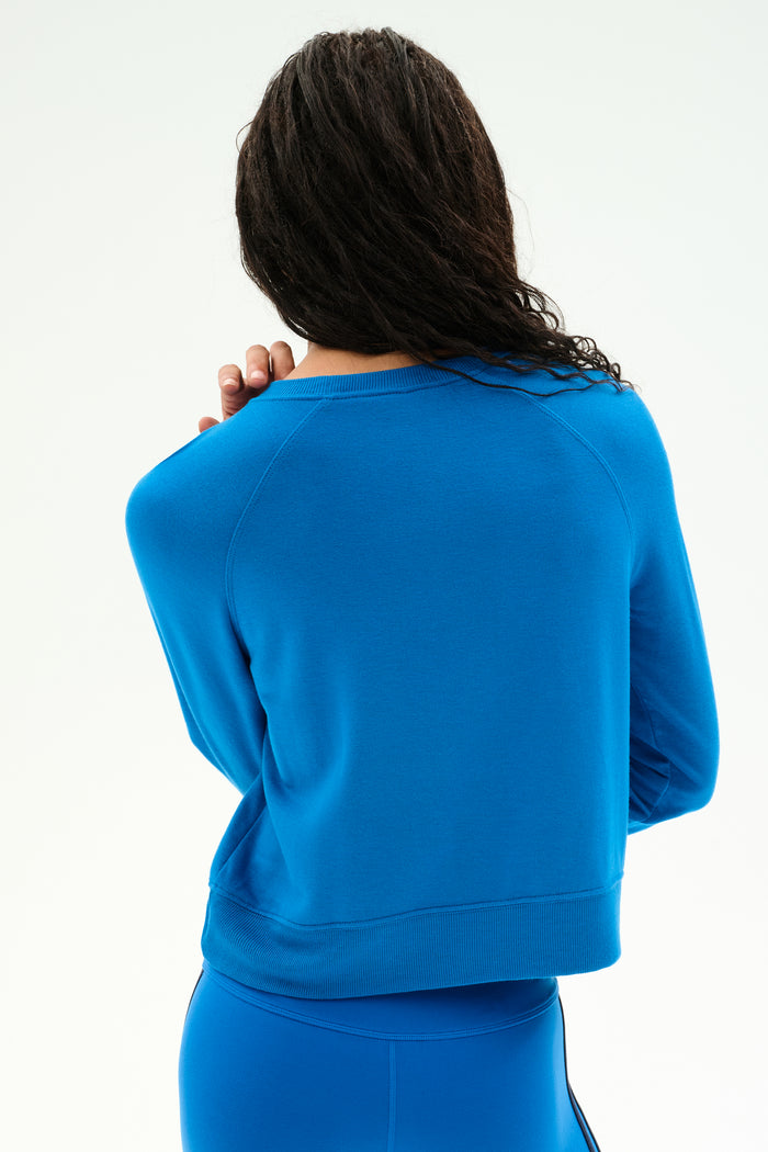Model with long dark hair shown from the back against a plain light background, wearing the SPLITS59 Brooks Fleece Long Sleeve Sweatshirt in Classic Blue with matching modal spandex pants.