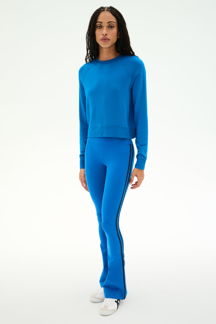 A person stands facing forward, wearing the SPLITS59 Brooks Fleece Long Sleeve Sweatshirt in Classic Blue paired with matching blue pants featuring black side stripes, crafted from a comfortable modal spandex blend, and white sneakers, against a plain white background.