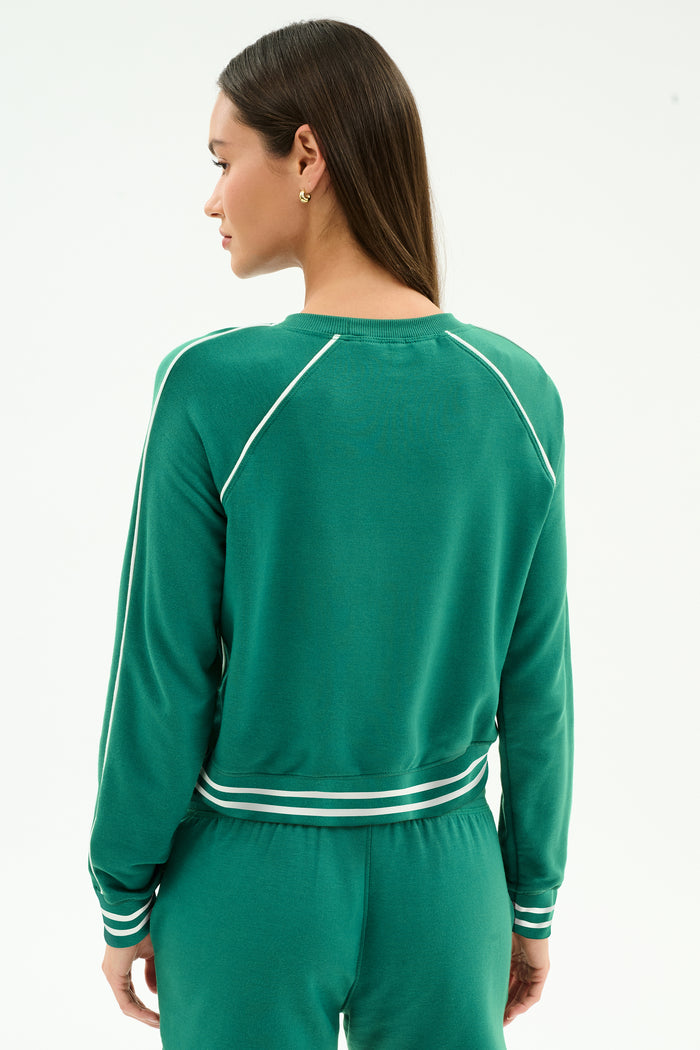 A woman with long straight hair and gold stud earrings wears the SPLITS59 Brooks Fleece Sweatshirt With Stripe in Arugula/White, featuring soft modal fabric and white stripes on the sleeves, cuffs, and waist.