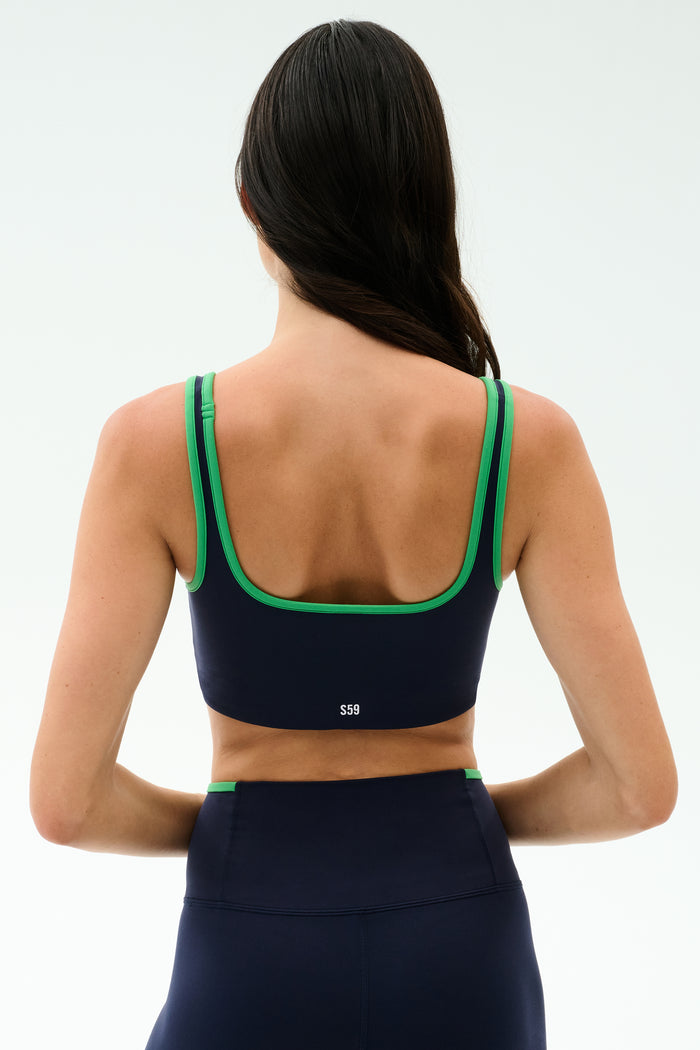 A woman with long dark hair wears the SPLITS59 Cait Rigor Bra in Indigo/Grass, featuring green trim and matching high-waisted leggings made from Rigor fabric, shown from the back against a plain light background.