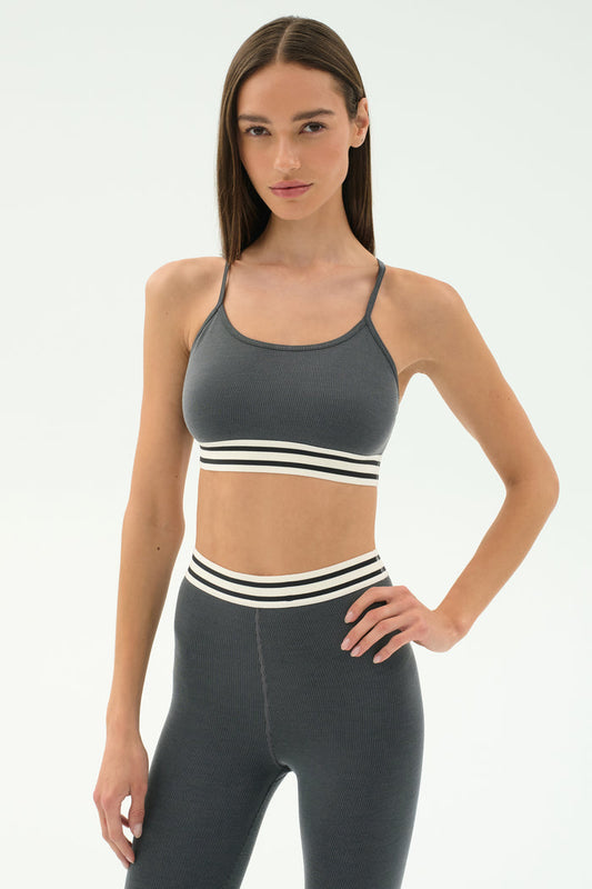 A woman in the SPLITS59 Cali Active Rib Bra in Heather Grey stands against a plain light background, paired with matching high-waisted leggings featuring white and black stripes.