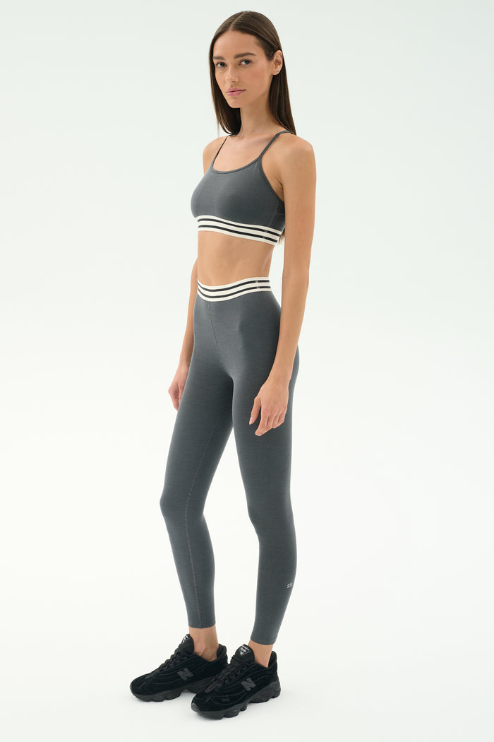 A woman stands against a plain background wearing the SPLITS59 Cali Active Rib Bra in Heather Grey with matching leggings featuring white striped bands, and black sneakers.