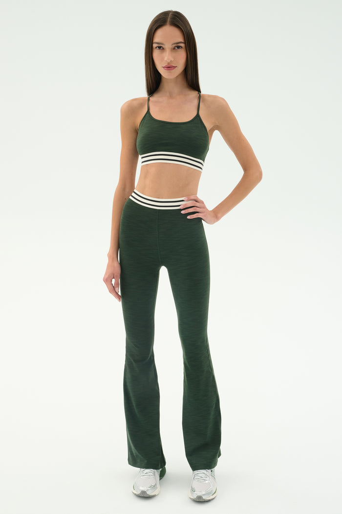 A woman stands in a studio wearing the SPLITS59 Cali Active Rib Bra in Heather Military with matching high-waisted leggings featuring white stripes, paired with white sneakers.