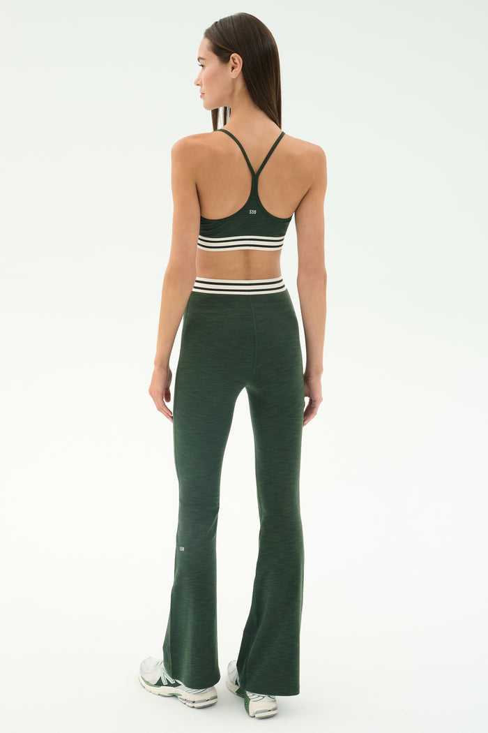 A woman stands facing away in the SPLITS59 Cali Active Rib Bra - Heather Military, paired with matching flared leggings featuring white stripes and white sneakers, set against a plain light background.