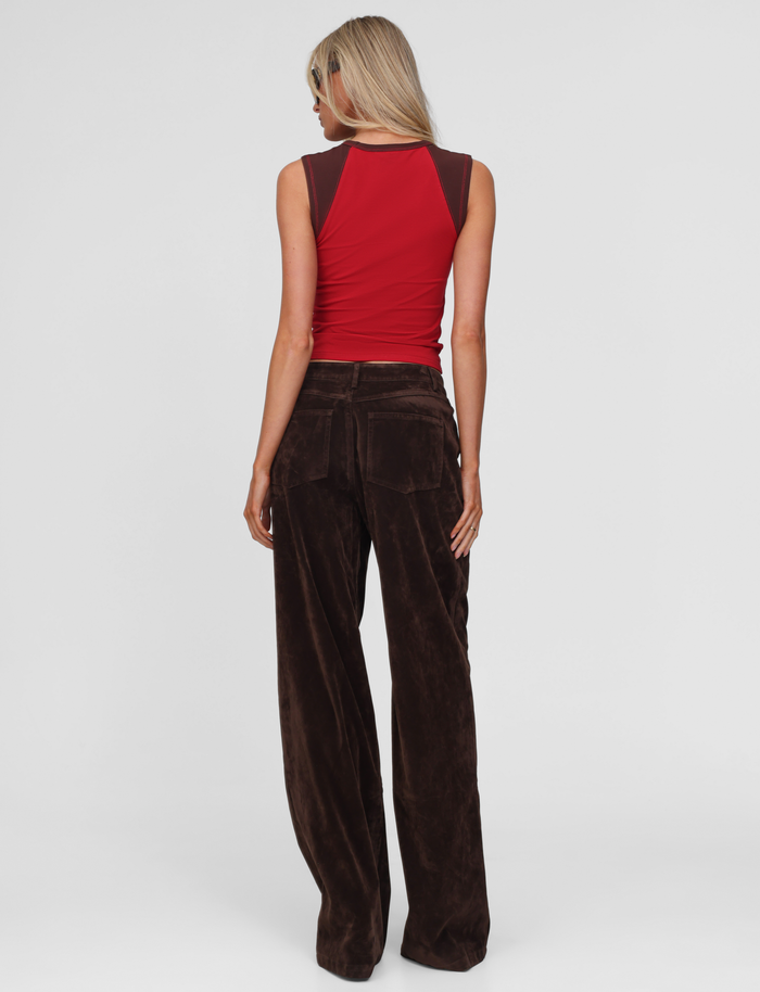 A woman with long blonde hair stands facing away on a white background, wearing TIGERMIST’s CAMELLIA PANT in chocolate suede, a sleeveless red top with brown shoulders, and sunglasses.