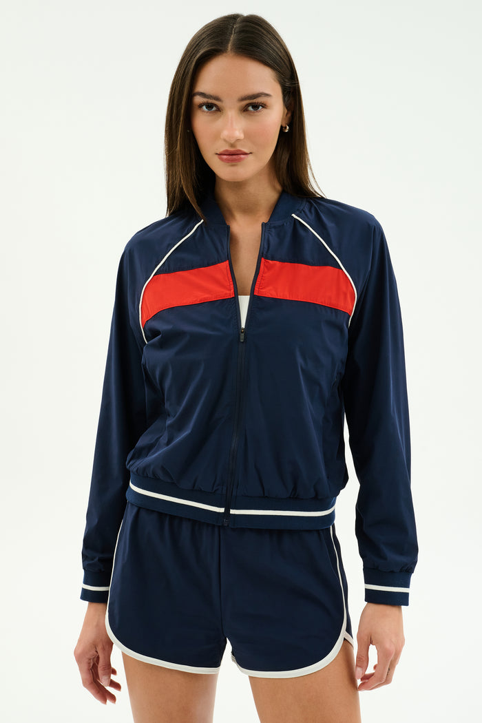 A woman faces forward wearing the SPLITS59 Carrie Woven Nylon Jacket in Indigo/Lotus Red, paired with matching navy blue spandex shorts featuring white trim.