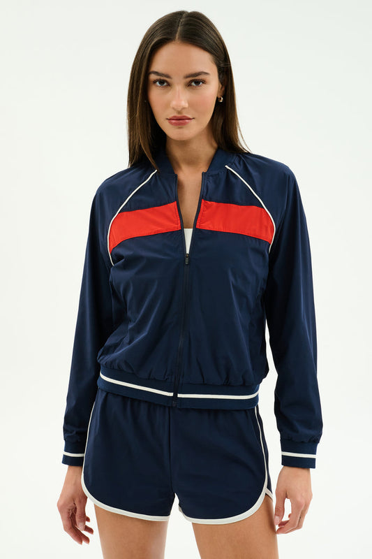 A woman faces forward wearing the SPLITS59 Carrie Woven Nylon Jacket in Indigo/Lotus Red, paired with matching navy blue spandex shorts featuring white trim.