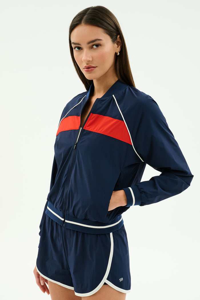 A woman wears the SPLITS59 Carrie Woven Nylon Jacket in Indigo/Lotus Red with matching nylon shorts featuring white trim, standing with one hand in her pocket.