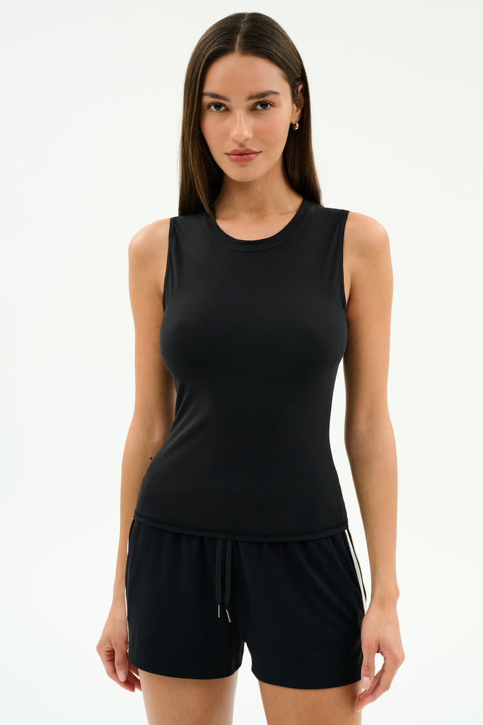 A woman stands against a plain background, wearing the SPLITS59 Carter Airweight Lite Tank in black and black spandex shorts.
