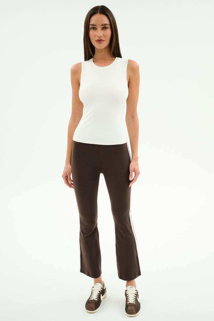 A woman faces forward against a white background, wearing the SPLITS59 Carter Airweight Lite Tank in white, paired with brown flared spandex pants with side stripes and brown sneakers.