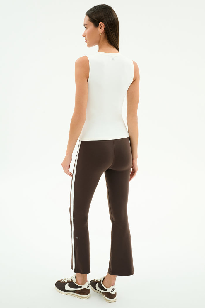 A woman faces away against a white background, wearing the SPLITS59 Carter Airweight Lite Tank in white, brown flared pants with side stripes, and brown sneakers.
