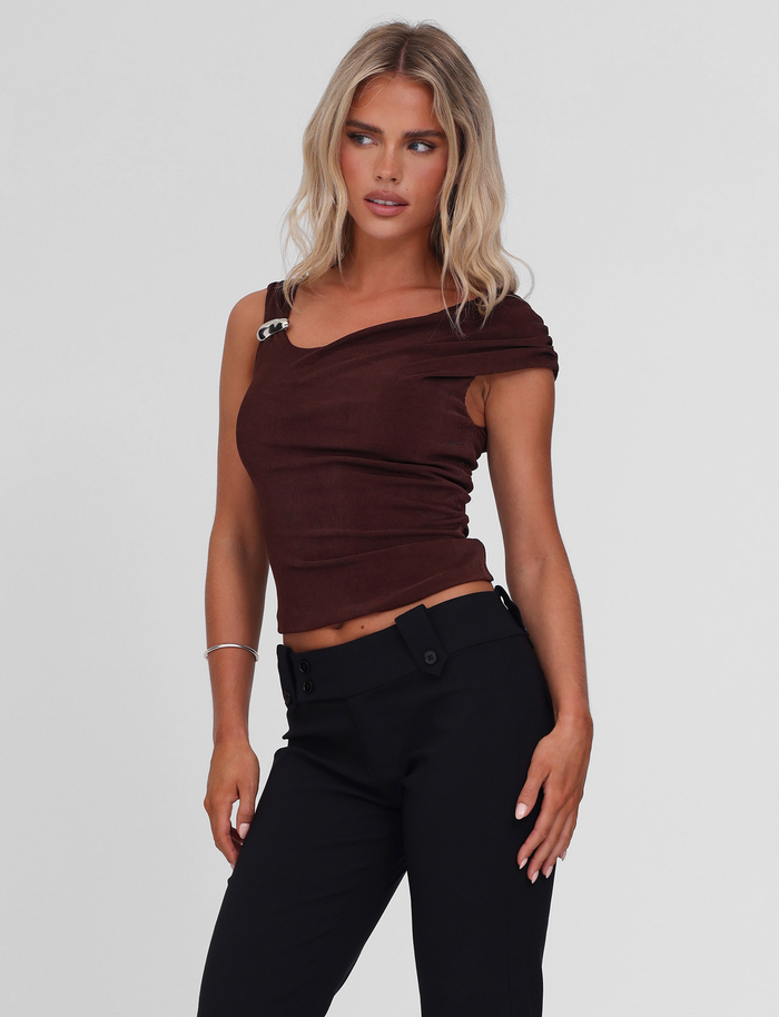 A woman with long blonde hair models the TIGERMIST CASSIAN TOP in chocolate, a slinky jersey one-shoulder style with metal detail, paired with black pants and posing against a plain light background.
