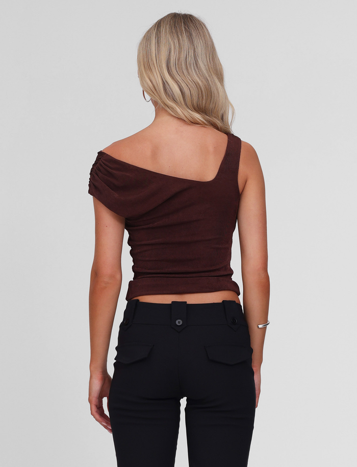 A woman with long blonde hair is shown from the back wearing the TIGERMIST CASSIAN TOP in chocolate and black pants with buttoned back pockets, standing against a plain white background.