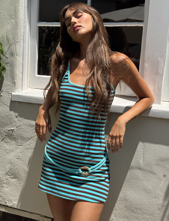 A woman with long brown hair stands outside in sunlight, wearing the TIGERMIST CELESTE MINI DRESS - BLUE STRIPE featuring a belted waist, adjustable shoulder straps, and a knotted hem, posing with eyes closed by a white wall and window.