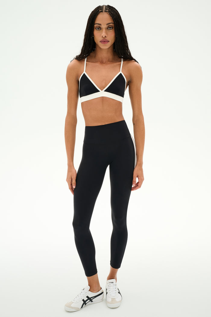A woman stands in a studio against a plain white background, wearing the SPLITS59 Clare High Waist Rigor 7/8 leggings in Black/White with a black sports bra featuring white straps and white sneakers.