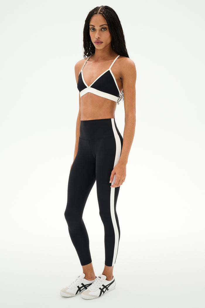 A person is wearing the SPLITS59 Clare High Waist Rigor 7/8 leggings in Black/White, crafted from Rigor fabric and featuring white side stripes, paired with a black sports bra and white sneakers—an ideal look for gym workouts. They are standing against a plain white background.