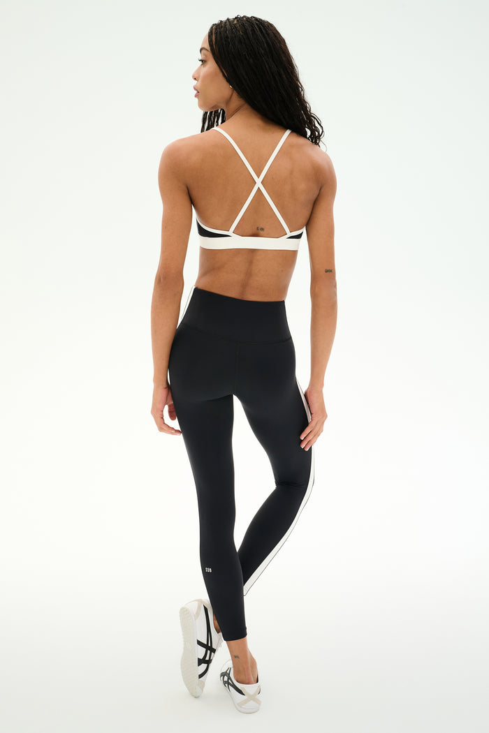A woman wearing the SPLITS59 Clare High Waist Rigor 7/8 leggings in Black/White and a white sports bra stands on one leg, facing away. The plain white background makes this an ideal inspiration for gym workouts.