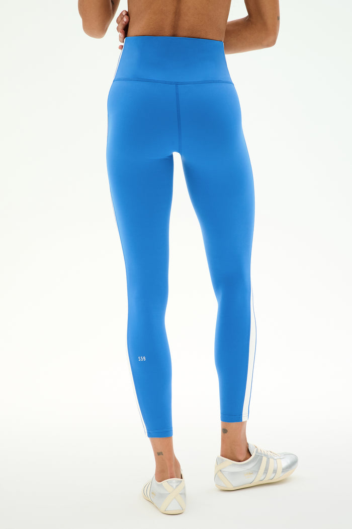 A person is shown from the back against a light background, wearing SPLITS59’s Clare High Waist Rigor 7/8 leggings in Classic Blue/White, made from a smooth nylon-spandex fabric, paired with white athletic shoes.