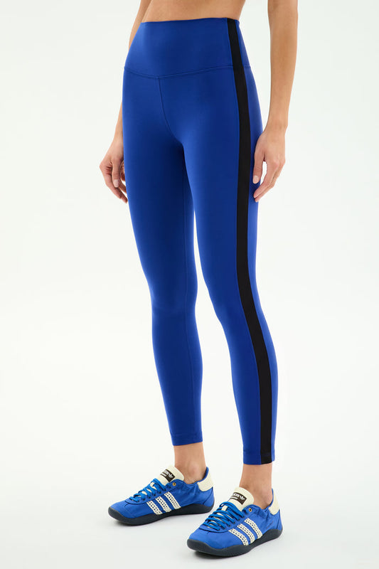 A person wears the SPLITS59 Clare High Waist Rigor 7/8 leggings in Cobalt/Black with blue Adidas sneakers, standing against a plain white background—ideal for gym workouts.