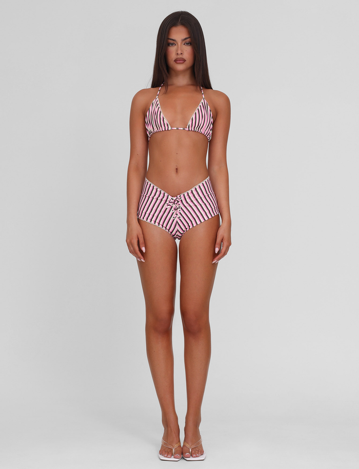 A woman with long dark hair and a neutral expression faces forward against a light gray background, wearing the TIGERMIST DAZZLE BIKINI BOTTOM - STRIPE and clear sandals.