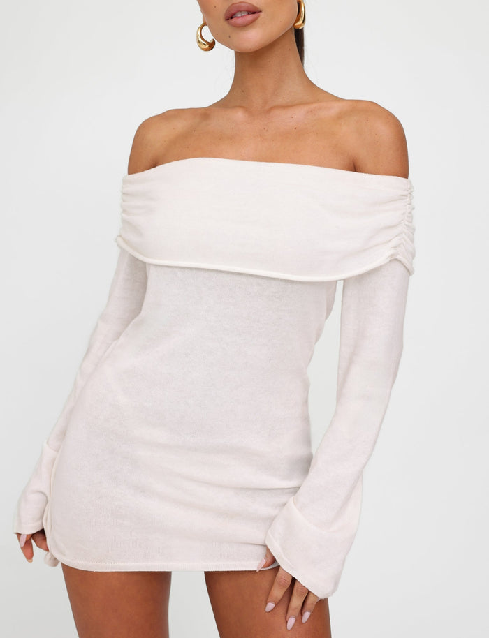 A woman wearing the TIGERMIST DIEDRE JUMPER DRESS - WHITE : CREAM, an off-the-shoulder, long-sleeve ivory mini dress, poses against a plain white background. The dress boasts a folded, draped neckline and slightly flared sleeves. She completes her look with large gold hoop earrings.