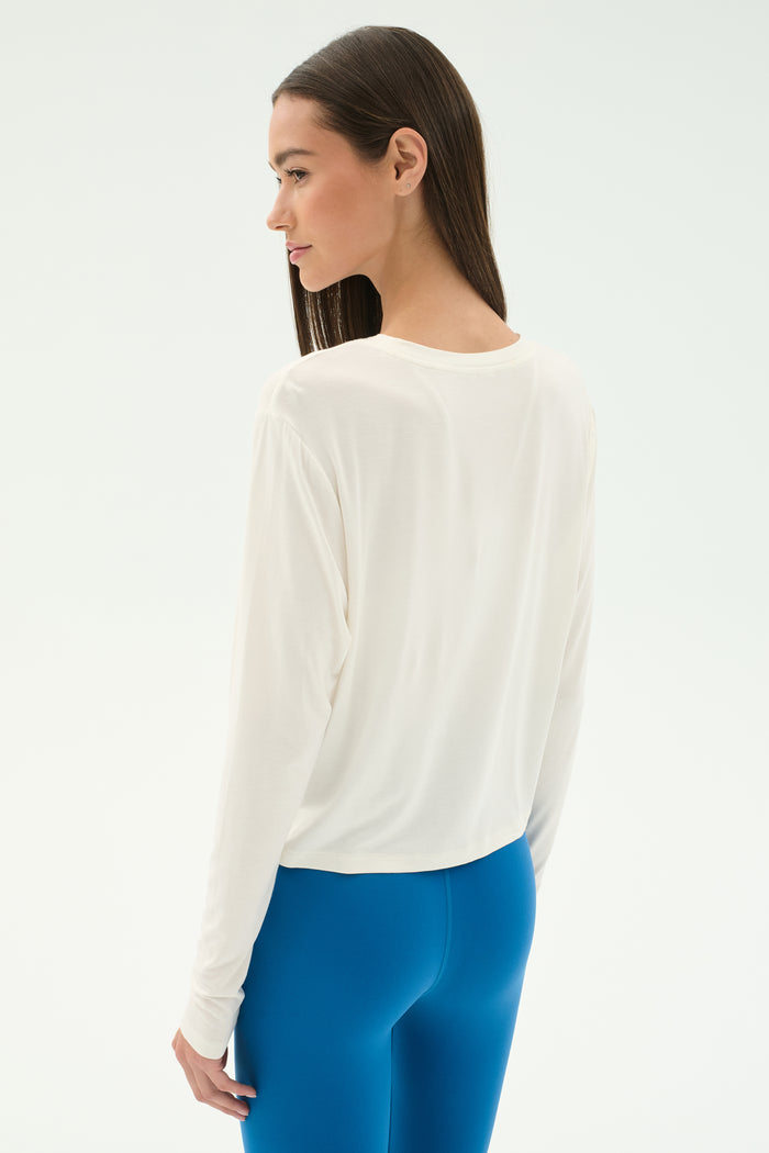 A woman stands sideways against a light background, wearing the SPLITS59 Djuna Long Sleeve in white and bright blue leggings.
