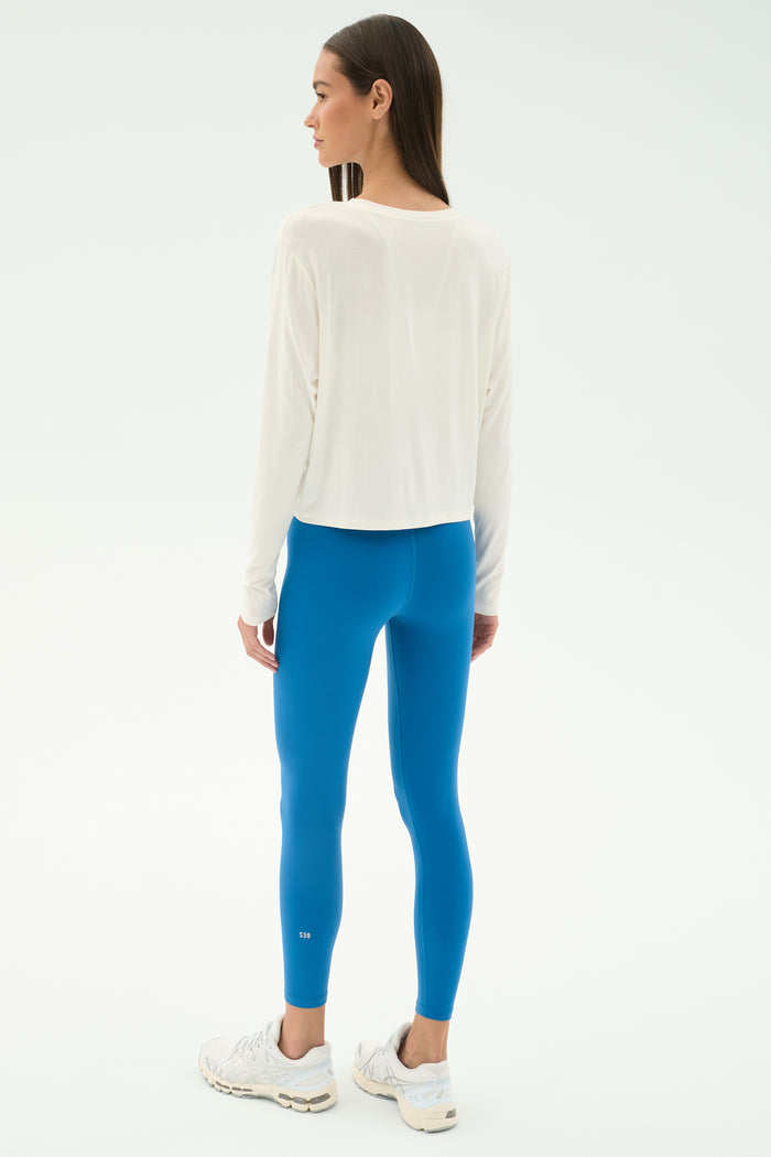 A woman faces away, wearing the SPLITS59 Djuna Long Sleeve - White, bright blue leggings, and white sneakers against a plain background—ideal for gym or outdoor workouts.