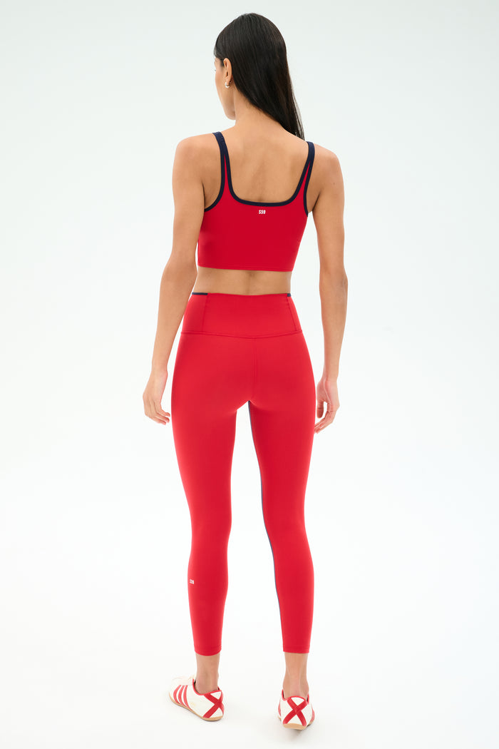 A woman faces away, wearing SPLITS59 Easton Rigor High Waist 7/8 leggings in Pirate Red/Indigo and a matching sports bra, paired with red-and-white athletic sandals against a plain light background.