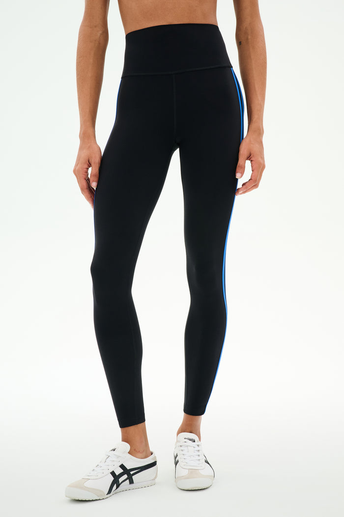 A person wearing SPLITS59 Ella High Waist Airweight 7/8 leggings in Black/Classic Blue, paired with white sneakers, standing against a plain white background.
