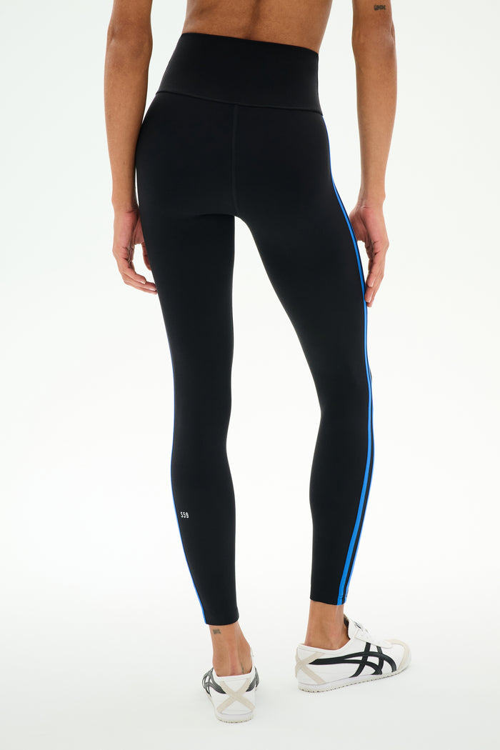 A person wearing SPLITS59 Ella High Waist Airweight 7/8 leggings in Black/Classic Blue and white sneakers stands against a plain light background.
