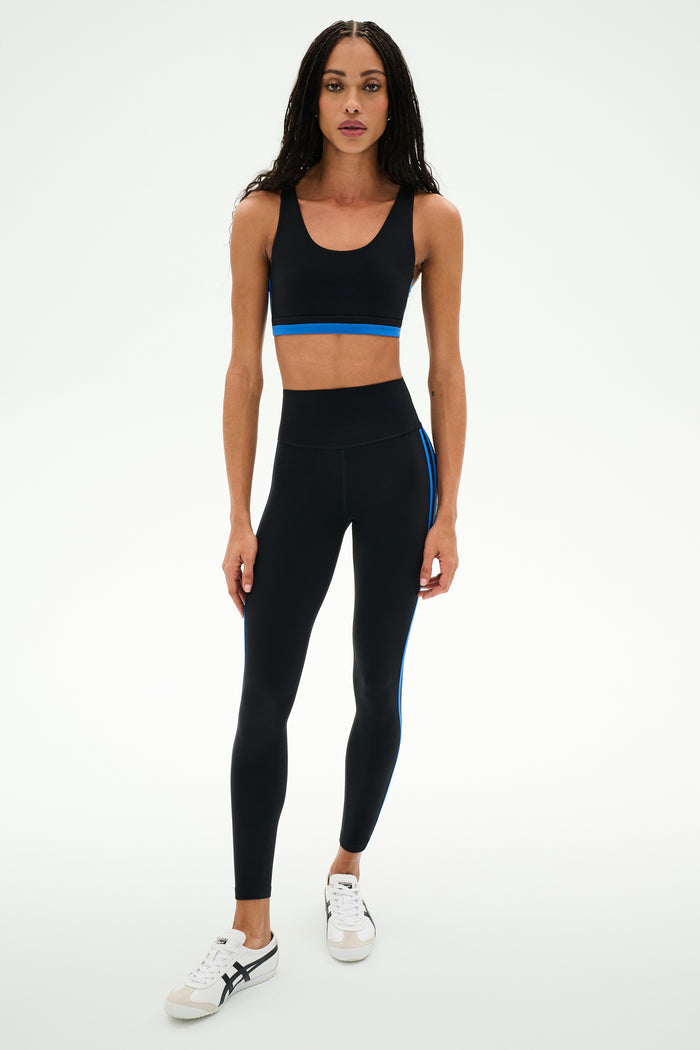 A person is wearing the SPLITS59 Ella High Waist Airweight 7/8 leggings in Black/Classic Blue, paired with a black sports bra and white athletic shoes, standing against a plain white background.
