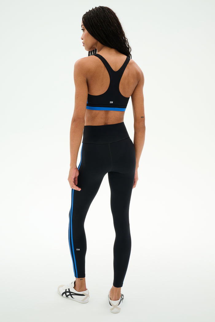 A person is standing on a white background, facing away from the camera, wearing SPLITS59's Ella High Waist Airweight 7/8 leggings in Black/Classic Blue, paired with a black sports bra and white athletic shoes.