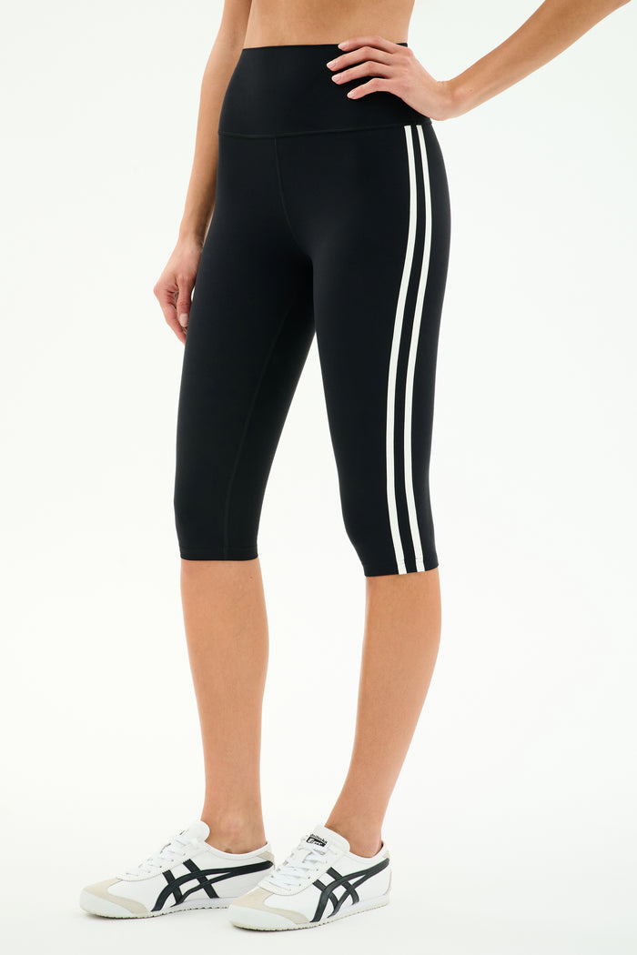A person wears SPLITS59 Ella High Waist Airweight Capri in Black/White, featuring two white side stripes, paired with white sneakers with black accents, standing against a plain white background.