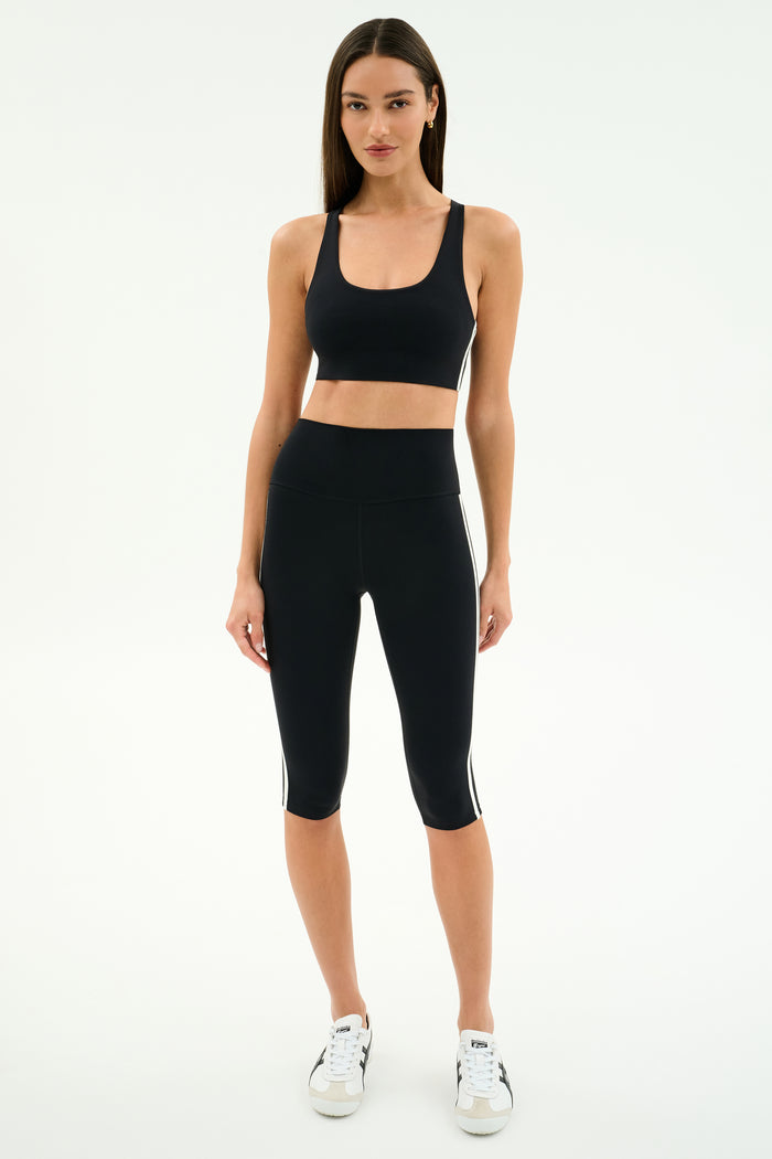 A woman stands against a plain background wearing the SPLITS59 Ella High Waist Airweight Capri in Black/White, a black sports bra, and white sneakers.